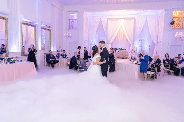 A bride and groom dancing at a wedding reception in a ballroom decorated with white drapery, floral centerpieces, and low-lying fog effects.
