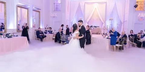 A bride and groom dancing at a wedding reception in a ballroom decorated with white drapery, floral centerpieces, and low-lying fog effects.