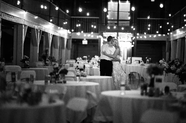 A black and white photograph of a bride and groom dancing at a wedding reception in a venue with string lights and round tables.