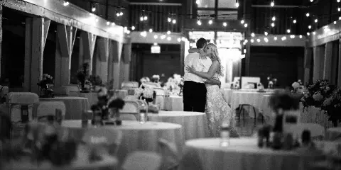 A black and white photograph of a bride and groom dancing at a wedding reception in a venue with string lights and round tables.