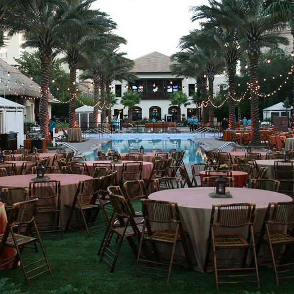 An outdoor event venue featuring round tables with pink linens, wooden chairs, and string lights hanging from palm trees near a swimming pool.