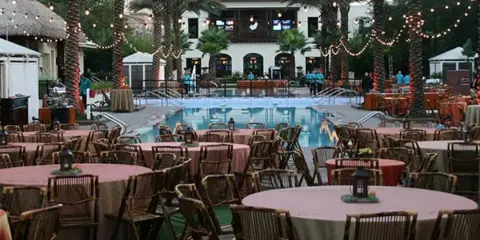 An outdoor event venue featuring round tables with pink linens, wooden chairs, and string lights hanging from palm trees near a swimming pool.