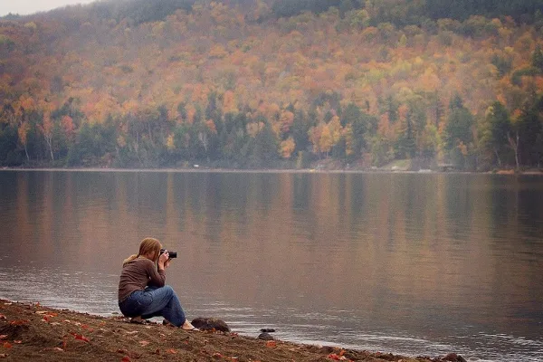 A photographer crouched by a lake shooting with a camera, with autumn forested hills in the background.