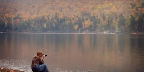 A photographer crouched by a lake shooting with a camera, with autumn forested hills in the background.