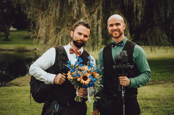 Two men in formal attire holding a wedding bouquet and a professional video camera with a gimbal stabilizer.