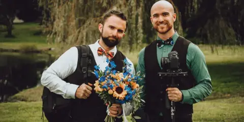 Two men in formal attire holding a wedding bouquet and a professional video camera with a gimbal stabilizer.