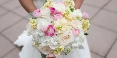 Bride in a white wedding dress holding a round bouquet of white and pink roses, ranunculus, and hydrangeas.