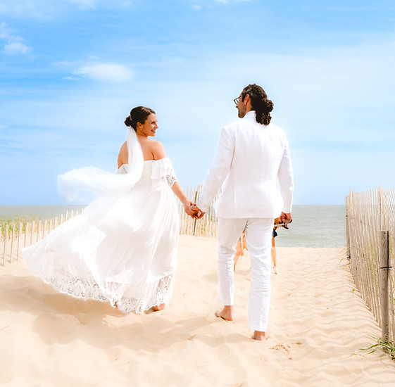 A bride and groom in white wedding attire walking hand-in-hand on a sandy beach near the ocean.