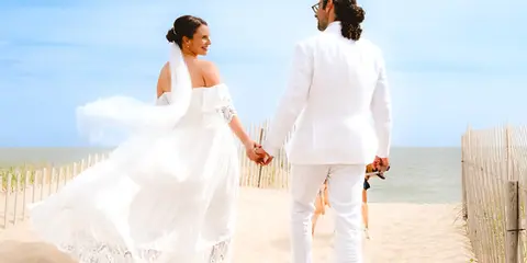 A bride and groom in white wedding attire walking hand-in-hand on a sandy beach near the ocean.