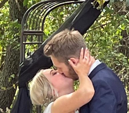 A bride and groom kissing under a black-draped outdoor ceremony arch surrounded by trees.