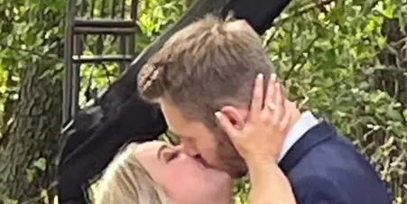 A bride and groom kissing under a black-draped outdoor ceremony arch surrounded by trees.