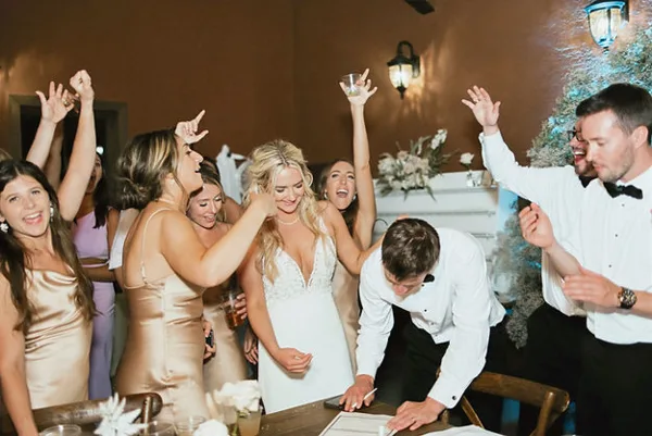 A bride and groom signing a document at a table while surrounded by cheering guests at a wedding reception.