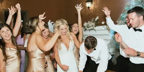 A bride and groom signing a document at a table while surrounded by cheering guests at a wedding reception.