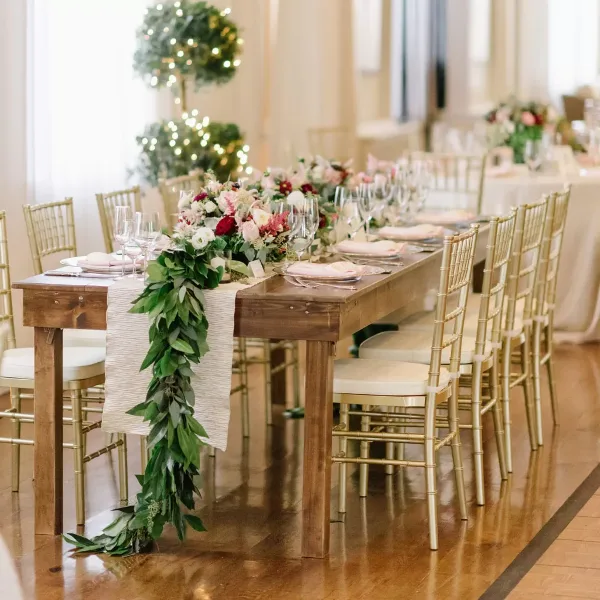 A long wooden dining table set with white linens and floral centerpieces, surrounded by gold Chiavari chairs in a bright event venue.