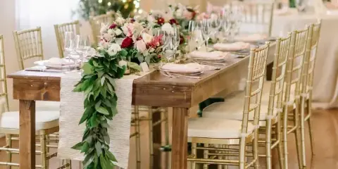A long wooden dining table set with white linens and floral centerpieces, surrounded by gold Chiavari chairs in a bright event venue.