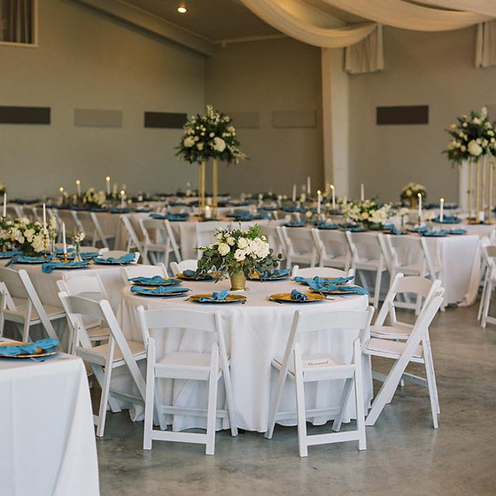 Indoor wedding reception room with round tables, white tablecloths, blue napkins, floral centerpieces, and white chairs.