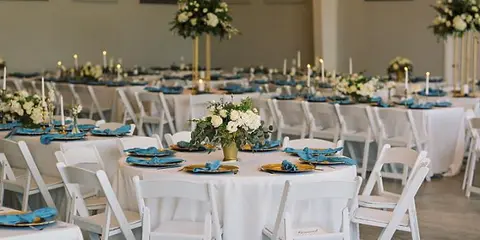 Indoor wedding reception room with round tables, white tablecloths, blue napkins, floral centerpieces, and white chairs.