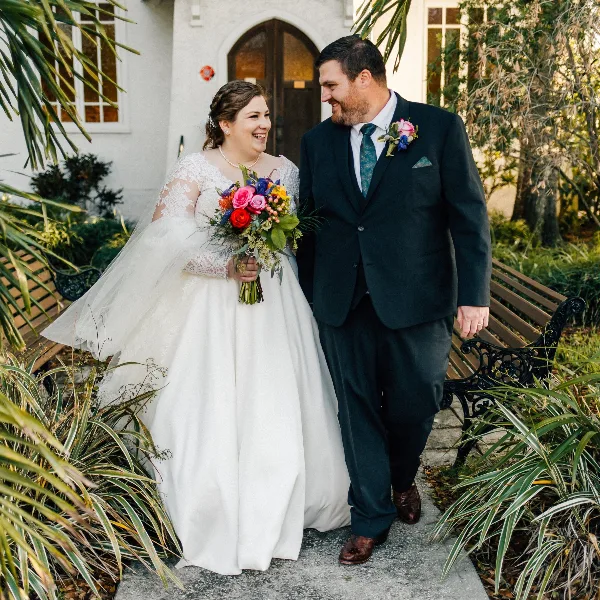 A bride and groom standing together outdoors in a garden setting, with the bride holding a colorful bouquet.