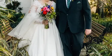 A bride and groom standing together outdoors in a garden setting, with the bride holding a colorful bouquet.