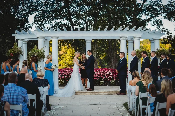 A wedding officiant standing between a bride and groom during a ceremony under a white pergola.