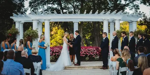 A wedding officiant standing between a bride and groom during a ceremony under a white pergola.