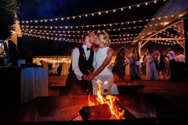 A bride and groom dancing at an outdoor wedding reception under string lights with a fire pit in the foreground.