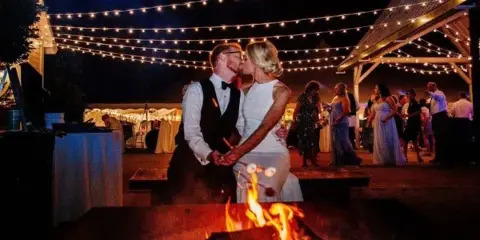 A bride and groom dancing at an outdoor wedding reception under string lights with a fire pit in the foreground.