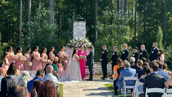 A wedding ceremony taking place outdoors with a bride, groom, and officiant standing before a stone altar, surrounded by a wedding party and seated guests.