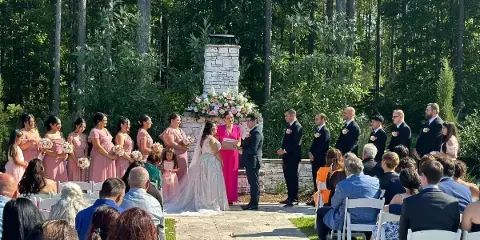 A wedding ceremony taking place outdoors with a bride, groom, and officiant standing before a stone altar, surrounded by a wedding party and seated guests.