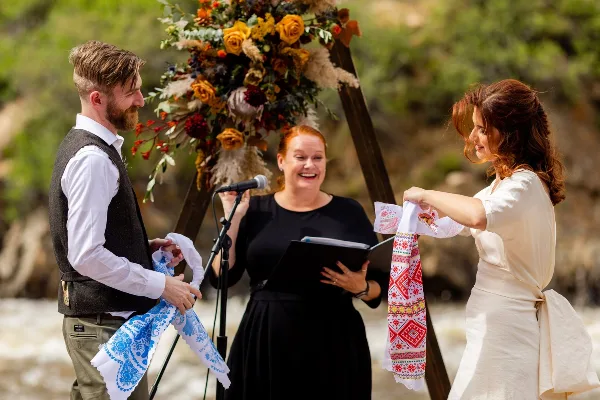 A female officiant leads a wedding ceremony where a couple participates in a unity ritual with embroidered sashes.