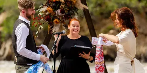A female officiant leads a wedding ceremony where a couple participates in a unity ritual with embroidered sashes.