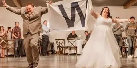A bride and groom dancing in a wedding reception hall, holding up a banner with the letter 'W'.