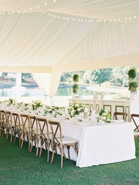 A long white-draped wedding table set with floral centerpieces and wooden chairs under a draped tent with string lights.