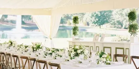 A long white-draped wedding table set with floral centerpieces and wooden chairs under a draped tent with string lights.