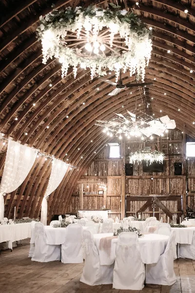 Rustic barn wedding reception interior with white-draped tables, floral chandeliers, and string lights under a wooden arched ceiling.