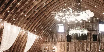 Rustic barn wedding reception interior with white-draped tables, floral chandeliers, and string lights under a wooden arched ceiling.