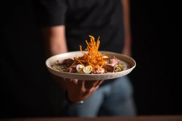 A person holding a white plate with a gourmet dish topped with shredded carrots and sliced eggs.