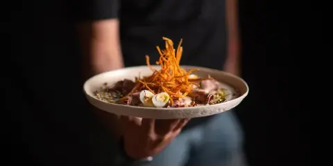 A person holding a white plate with a gourmet dish topped with shredded carrots and sliced eggs.