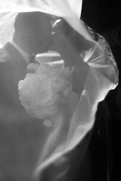 Black and white photograph of a bride and groom kissing under a veil with a bouquet of white roses.