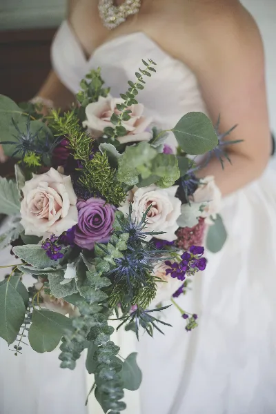 A bride in a white gown holding a cascading wedding bouquet featuring blush and purple roses, eucalyptus, and thistles.