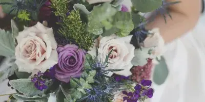 A bride in a white gown holding a cascading wedding bouquet featuring blush and purple roses, eucalyptus, and thistles.