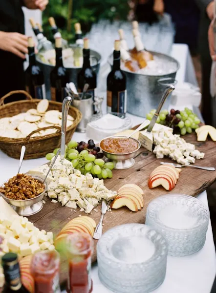 A catering table featuring a wooden board with assorted cheeses, grapes, nuts, and sliced apples, accompanied by wine bottles in ice buckets.