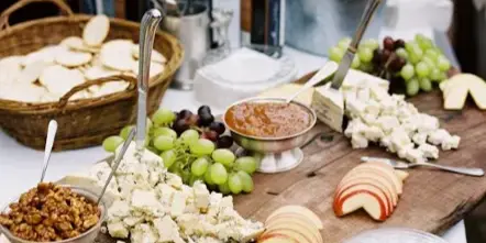 A catering table featuring a wooden board with assorted cheeses, grapes, nuts, and sliced apples, accompanied by wine bottles in ice buckets.