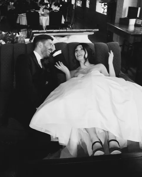 A black-and-white photo of a bride and groom laughing together on a couch at a wedding reception.