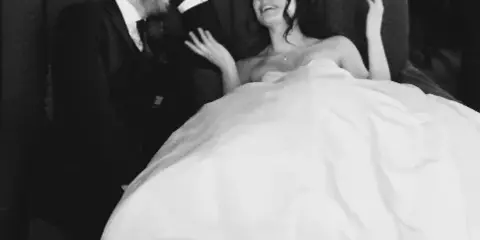 A black-and-white photo of a bride and groom laughing together on a couch at a wedding reception.