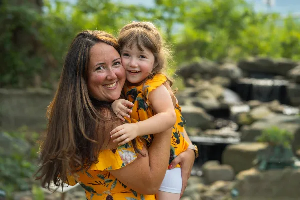 Mother and young daughter in matching yellow floral dresses smiling together outdoors near a rocky waterfall.