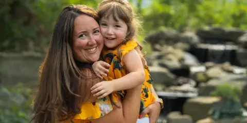 Mother and young daughter in matching yellow floral dresses smiling together outdoors near a rocky waterfall.