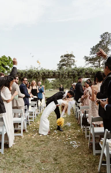 A bride and groom bowing to their guests during an outdoor wedding ceremony exit, with petals on the ground.