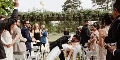 A bride and groom bowing to their guests during an outdoor wedding ceremony exit, with petals on the ground.