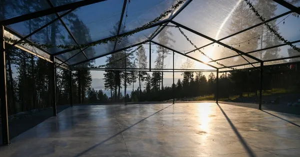 Open-sided glass wedding pavilion with concrete floor and string lights overlooking a lake and forest at sunset